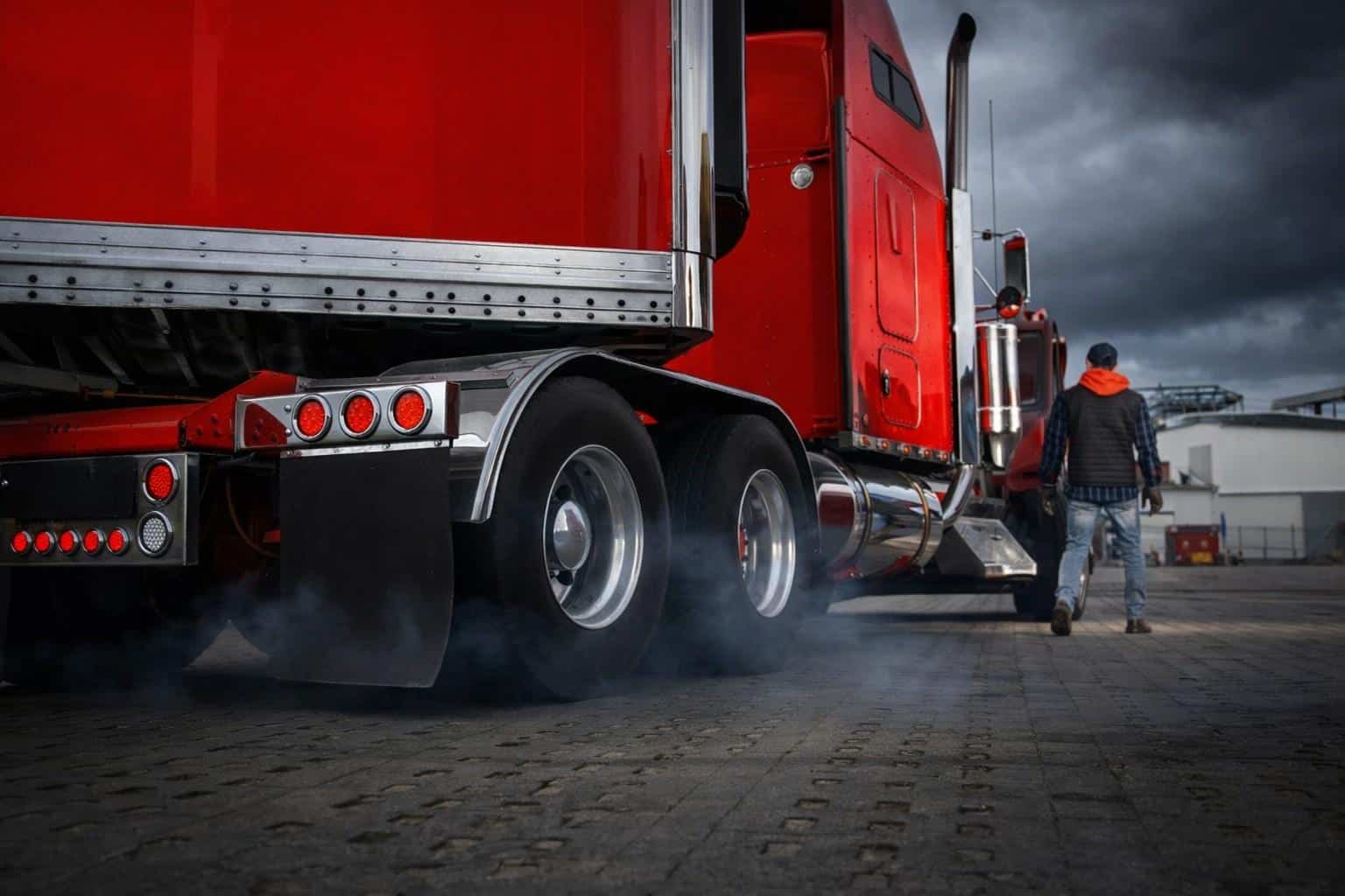 Red semi truck parked in an industrial yard with a driver walking beside the trailer under stormy skies