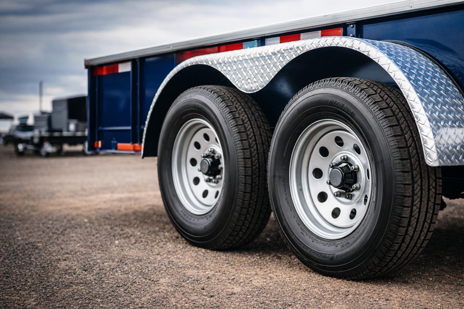 Close-up of dual-axle trailer tires with blue trailer frame and red reflectors on a gravel lot.