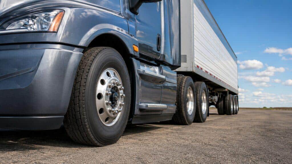 Close-up of a semi truck and trailer with strong focus on the front steer tire, drive tires, and trailer tires on an open paved lot under a blue sky.